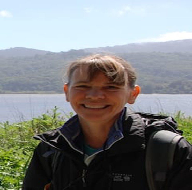 Smiling person outdoors with a scenic lake and hills in the background.