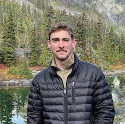 A man in a black puffer jacket standing by a lake with mountains and trees in the background.