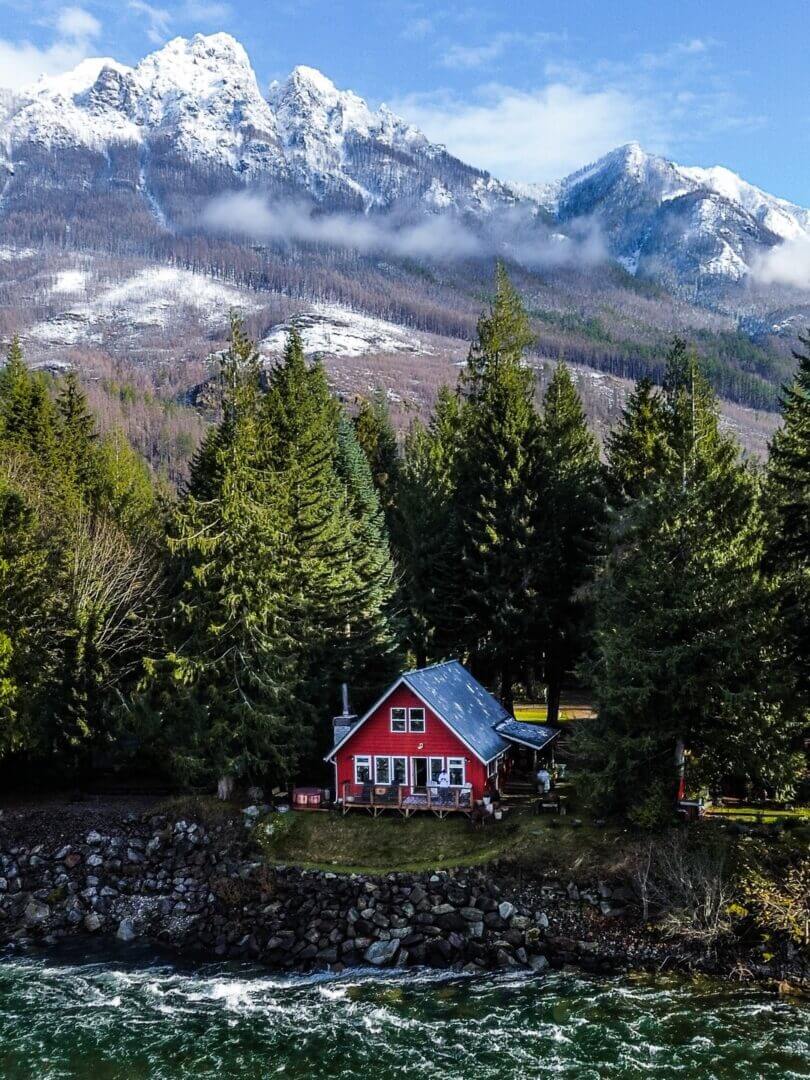 Red cabin nestled among pine trees with snowy mountains in the background.