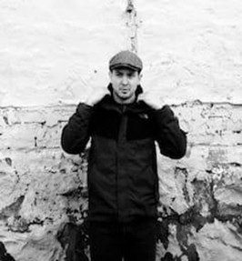 Man in a cap posing against a textured brick wall.