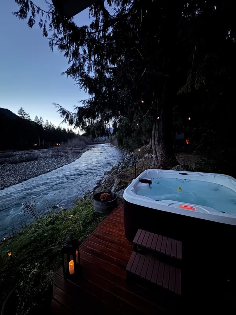 Outdoor hot tub beside a river at dusk with trees and mountains.