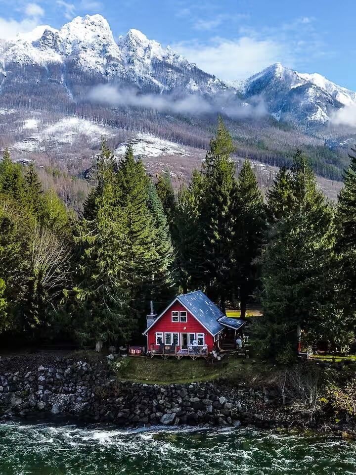 A red cabin nestled among pine trees with snowy mountains in the background.