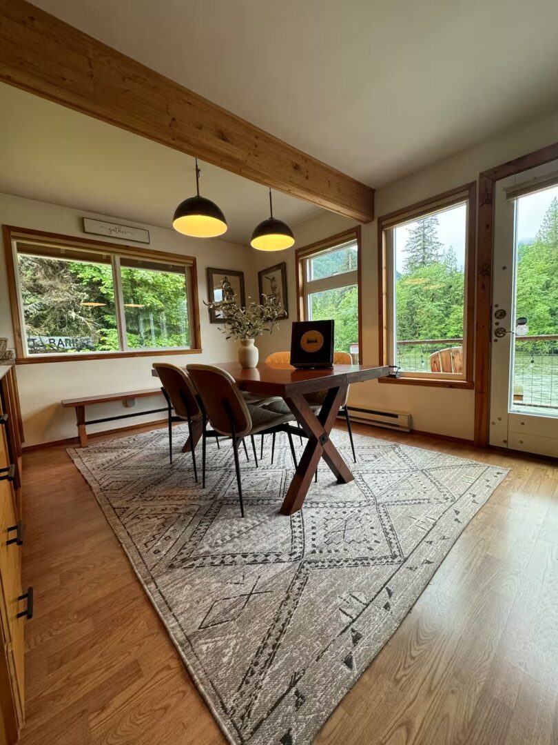 Bright dining room with wooden table and large windows overlooking greenery.