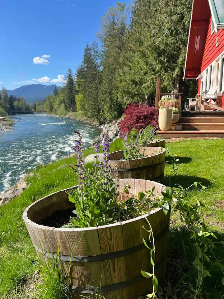 Riverfront garden with flowers in wooden barrels and mountain views.