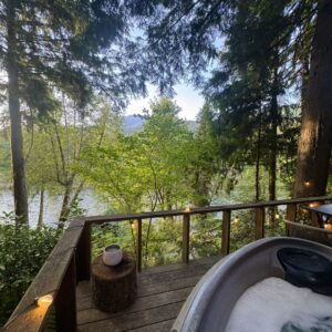 A serene lakeside view with a hot tub on a wooden deck surrounded by trees.