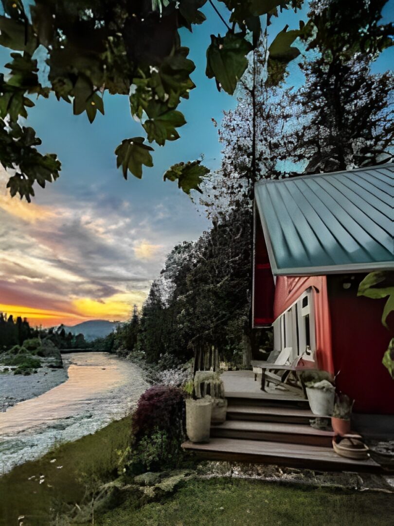 Cozy cabin by a river at sunset with vibrant sky and autumn leaves.