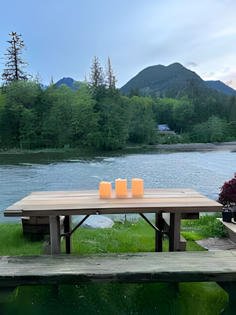 A wooden picnic table with candles by a river and mountains.