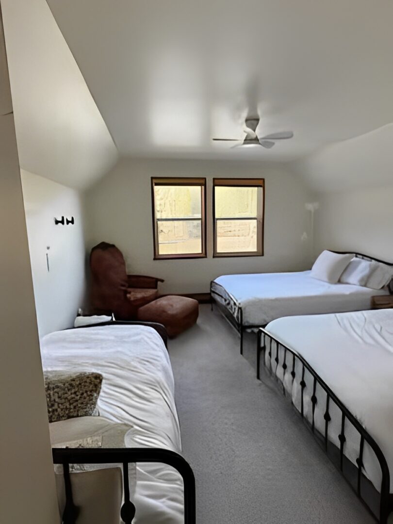 Cozy bedroom with two beds, an armchair, and a ceiling fan under sloped ceilings.