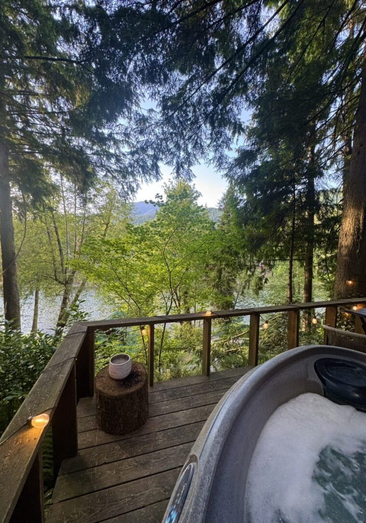 A serene lakeside view with a hot tub on a wooden deck surrounded by trees.